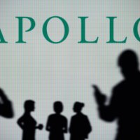 Apollo Global Management logo is seen on an LED screen in the background while a silhouetted person uses a smartphone