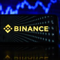 Binance logo displayed on a smartphone with stock market percentages on the background