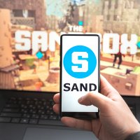 The Sandbox logo on a phone, which is in front of a laptop displaying the virtual world
