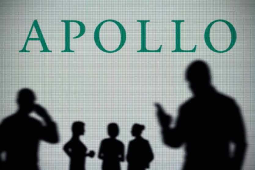 Apollo Global Management logo is seen on an LED screen in the background while a silhouetted person uses a smartphone