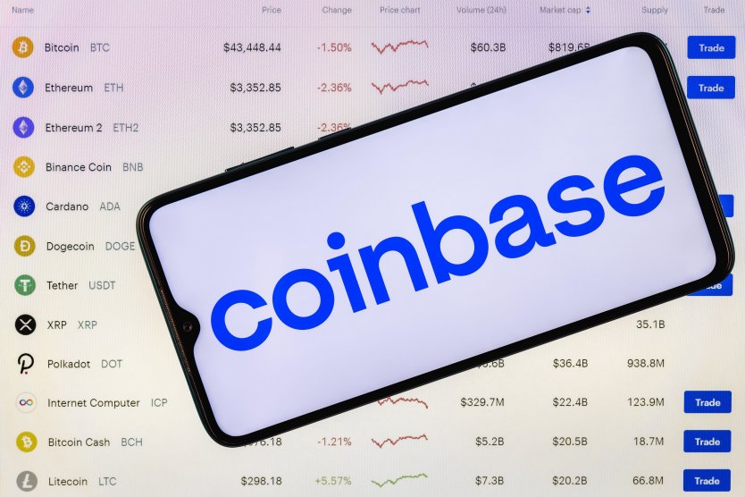 Coinbase logo on smartphone set against a computer screen showing list of cryptocurrencies traded on the exchange