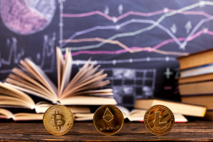 Crypto tokens stand on edge in front of graphs on a chalkboard and piles of books