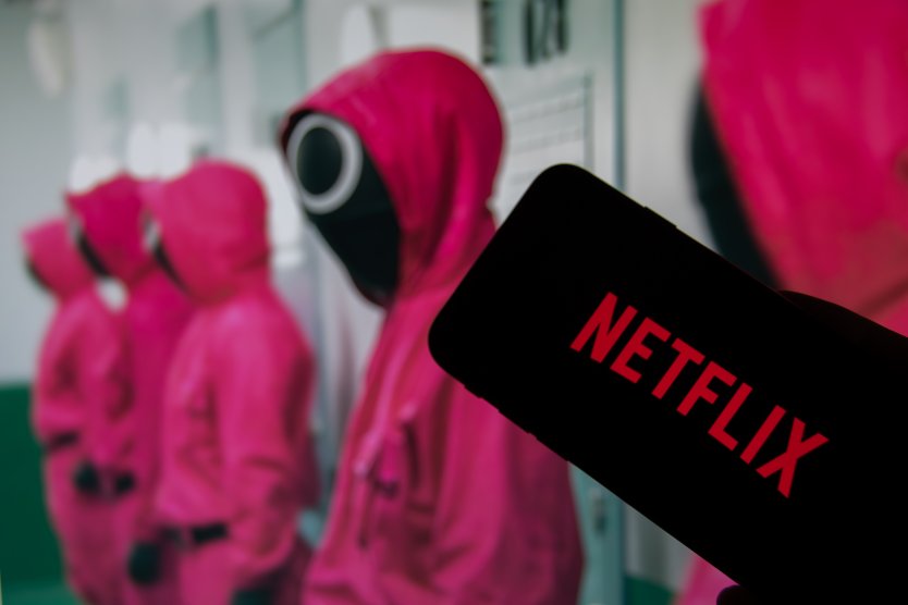The Netflix logo on the display of a smartphone in front of a television with the series Squid Game