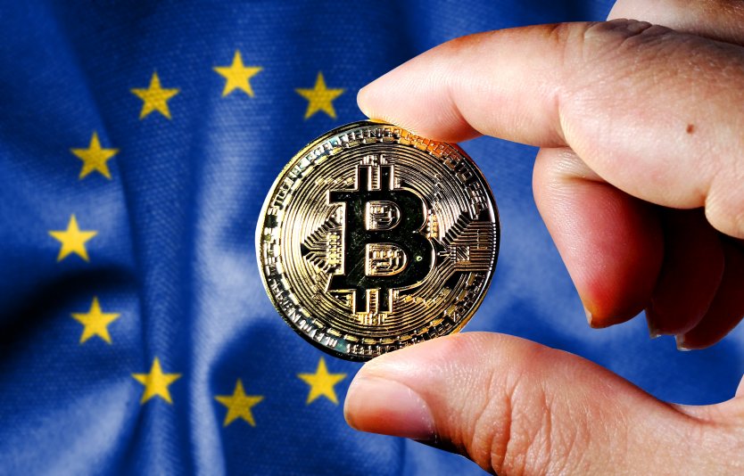 Hand holding a bitcoin, EU flag in background – Photo: Shutterstock